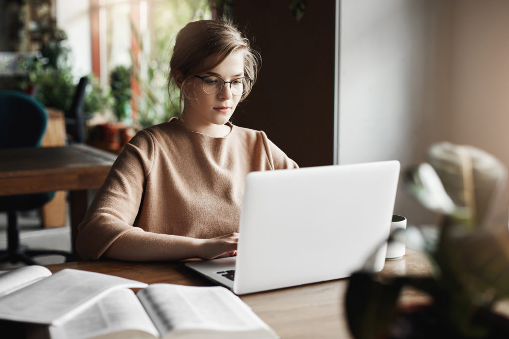 Work, lifestyle and business concept. Good-looking focused european female in trendy glasses sitting in cafe near laptop, working on notebook, surrounded with books, making notes for lesson with kids